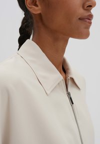 Beige Kragenjacke mit glatter Textur, auffälligem Reißverschluss und klassischem Kragendesign. Nahaufnahme des Schulterbereichs.