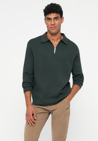 Mørkegrønn halv-zip genser med krage, lange ermer og ribbestrikkede mansjetter, sammen med lyse beige kordfløyelsbukser. Glatt tekstur.
