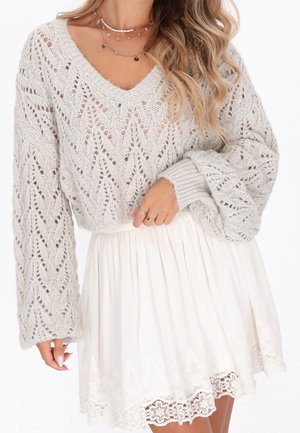 Femme portant un pull en maille gris clair avec des manches ballon, des colliers en or superposés et une jupe plissée blanche avec un bord en dentelle.