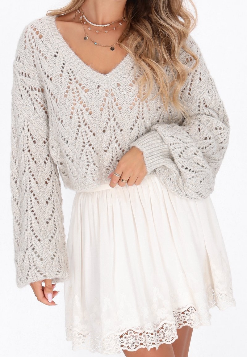 Femme portant un pull en maille gris clair avec des manches ballon, des colliers en or superposés et une jupe plissée blanche avec un bord en dentelle.