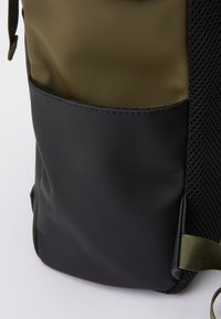 Sac à dos avec une section supérieure olive verte, un fond noir, un côté en mesh texturé, et une poche latérale, doté d'une sangle réglable et de coutures renforcées.