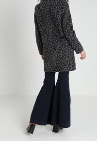 Kabát z vlny s leopardím potiskem v tmavě šedé barvě, s dlouhými rukávy, kombinovaný s rozšířenými tmavými džínovými kalhotami. Hladká textura a design střední délky.