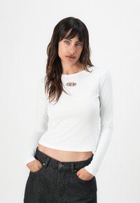 Hvit langermet kort topp med et sølvfarget ovalt smykke ved halsen, sammen med høy midje mørke denimbukser.