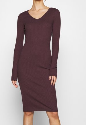 Kobieta w dopasowanej, prążkowanej sukience o długości do kolan, z długimi rękawami, w ciemnym śliwkowym kolorze z dekoltem w kształcie litery V, stojąca na jednolitym jasnym tle.