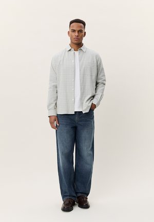 Άνδρας που στέκεται φορώντας ανοιχτό γκρι πουκάμισο με καρό μοτίβο και κουμπιά, λευκό μπλουζάκι, μπλε τζιν και σκούρα καφέ παπούτσια μπροστά σε απλό φόντο.