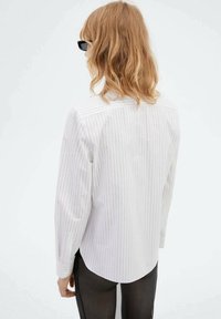 Persoon met schouderlang, rood haar, die zwarte zonnebril, een wit overhemd met streepjes en donkere broek draagt, staand tegen een witte achtergrond.