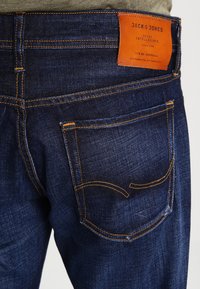 Vaqueros de mezclilla azul oscuro con costuras naranjas, una etiqueta de cuero con "JACK & JONES" y un único bolsillo trasero con un diseño curvado.
