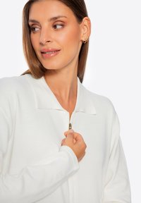 Chaqueta blanca con cremallera y cuello tipo polo, de textura acanalada. Presenta una cremallera dorada y un detalle bordado en la manga.