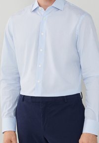 Chemise élégante à manches longues en tissu bleu clair à carreaux, avec col boutonné, fermeture à boutons sur le devant et boutons blancs. Portée avec un pantalon bleu foncé.