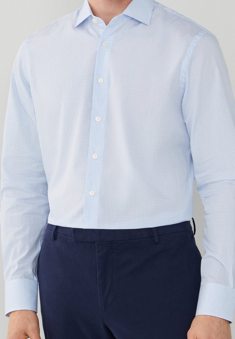Chemise élégante à manches longues en tissu bleu clair à carreaux, avec col boutonné, fermeture à boutons sur le devant et boutons blancs. Portée avec un pantalon bleu foncé.