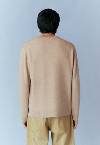 Pull en maille beige à manches longues, col rond et texture subtile, associé à un pantalon clair avec des coutures apparentes.