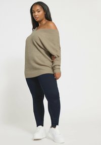 Beige off-the-shoulder ribbed neule paita yhdistettynä tummiin slim-fit farkkuihin ja valkoisiin lenkkareihin, jotka korostavat rentoa istuvuutta ja teksturoitua kangasta.