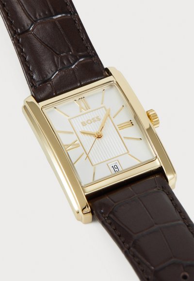 Montre rectangulaire dorée avec un cadran texturé blanc, un bracelet en cuir marron au motif crocodile et un affichage de la date à la position 6 heures.
