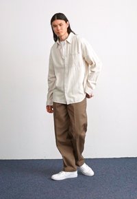 Hellgestreiftes, knöpfbares Hemd mit einer Tasche, kombiniert mit lockeren, beige-farbenen Hosen und weißen Turnschuhen; einfarbigem Hintergrund und blauem Teppich.
