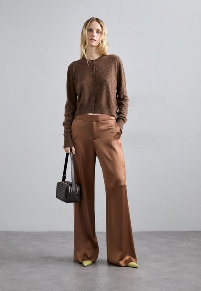Καφέ πλεχτό ζακέτα με υφαντή υφή, σε συνδυασμό με φαρδιά σατέν παντελόνια, με απαλές κίτρινες λεπτομέρειες στα παπούτσια και μια μικρή μαύρη τσάντα.