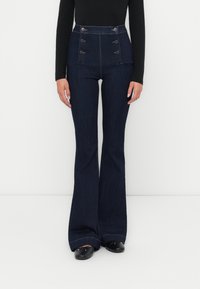 Pantalon évasé en denim foncé taille haute avec une fermeture à boutons, deux poches latérales et un ourlet effiloché. Le matériau semble doux avec un léger éclat.