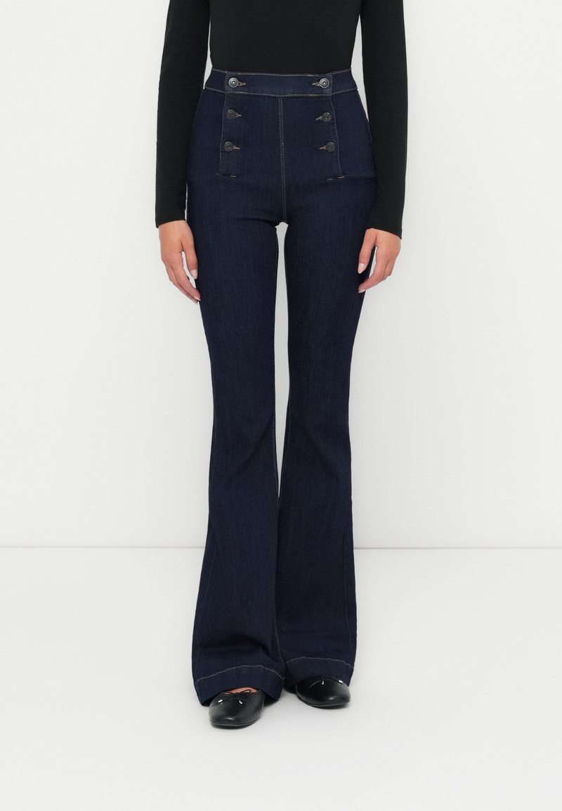 Pantalon évasé en denim foncé taille haute avec une fermeture à boutons, deux poches latérales et un ourlet effiloché. Le matériau semble doux avec un léger éclat.