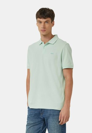 Młody mężczyzna ubrany w jasnozieloną koszulkę polo z krótkim rękawem i niebieskie dżinsy, stojący na tle jednolitego białego tła.