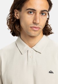 Lys beige polo shirt lavet af tekstureret stof. Har en krave, tre knapper og et lille mørkt logo på brystet.