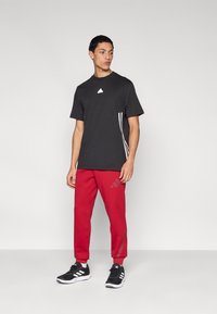 Μαύρο μπλουζάκι με λευκό λογότυπο και τρεις λευκές ρίγες στο πλάι, συνδυασμένο με κόκκινο παντελόνι που διαθέτει διακριτικό λογότυπο και ελαστικές μανσέτες, φορεμένο με μαύρα παπούτσια.