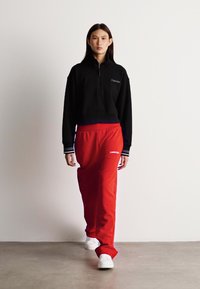 Black zip-up sweatshirt with a fitted hem paired with loose red sweatpants. White shoes. Sweatpants feature a logo detail on the left thigh.
