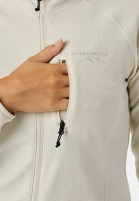 Light beige fleece jacket with a zippered pocket on the chest, featuring a pull tab and subtle brand logo embroidered on the fabric.
