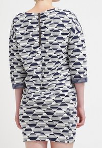 Femme portant une robe à motif géométrique bleu marine et blanc, manches trois-quarts, avec une fermeture éclair apparente dans le dos.