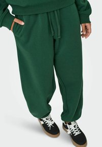 Grønne sweatpants med løs pasform, der har sidelommer og snøre i taljen, parret med sorte sneakers med hvide detaljer.