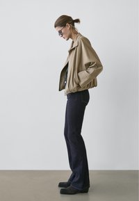 Beige bomberjacka med sänkta axlar, svart topp, mörka utsvängda jeans och ankelboots. Slät tyg, avslappnad passform, minimalistisk design.