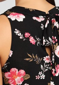 Schwarzes Blumentop mit rosa und weißen Blumen, einem hohen Kragen mit einer Schleife am Rücken und einem floralen Muster auf weichem Stoff.