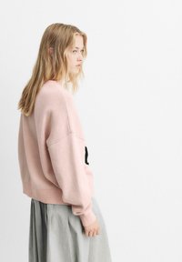 Maglione a maglia rosa chiaro con vestibilità rilassata e spalle scese, abbinato a una gonna grigia fluida, con un dettaglio di design nero sulla parte anteriore.