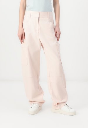 Femme portant un pantalon cargo à jambes larges rose clair avec poches avant et des baskets blanches, debout sur un fond blanc uni.