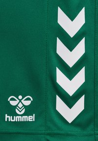 Camiseta deportiva verde con un patrón de chevrón blanco en el costado, que presenta un logotipo redondeado y el texto "hummel" en blanco en la parte inferior. Textura de tejido suave.