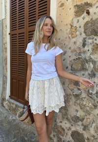 T-shirt blanc à manches courtes associé à une jupe en dentelle crème, fluide et à plusieurs couches. Un sac en osier ajoute une touche texturée. Mur en pierre en arrière-plan.