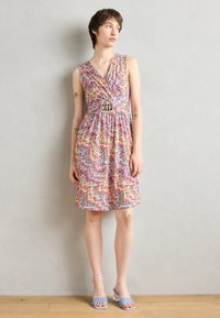 Robe sans manches colorée avec un motif en zigzag, une taille cintrée et un détail de ceinture métallique. Porte des chaussures bleu clair. Fond uni.