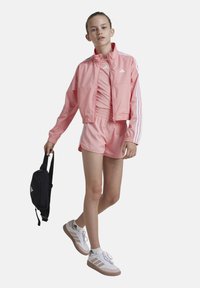 Chaqueta deportiva rosa con rayas blancas, pantalones cortos a juego y zapatillas blancas con acentos beige; llevando un pequeño bolso negro.
