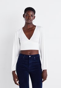 Biely dlhý rukáv crop top s hlbokým V-krkom a nariasením v oblasti pásu, v kombinácii s tmavomodrými zamatovými nohavicami s vysokým pásom.