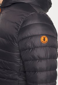 Giubbotto puffa nero con design trapuntato orizzontale. Caratterizzato da un logo circolare arancione sulla manica. Il materiale appare leggero e lucido.