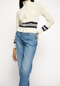 Femme portant un pull zippé crème avec des rayures noires et un jean en denim bleu clair, debout avec une main sur la hanche.