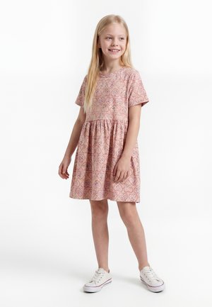 Ung pige med langt blondt hår iført en lyserød mønstret kortærmet kjole og hvide sneakers, stående og let smilende til siden.