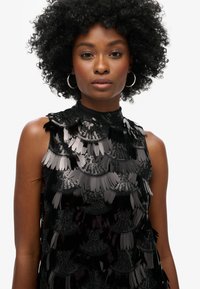 Haut sans manches noir avec un col haut, orné de franges superposées et de paillettes, créant un effet texturé et scintillant.