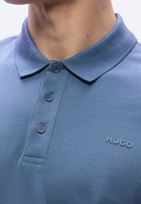 Gros plan d’une personne portant un polo bleu à col avec trois boutons et un petit logo en relief "HUGO" sur la poitrine.