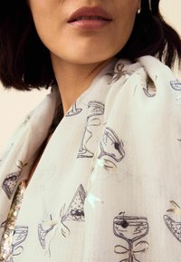 Viegls šalle, kas izgatavota no caurspīdīga balta auduma ar atkārtotu kokteiļu glāžu, loku un konfekšu rakstu ar zilu un zeltītu akcentu.