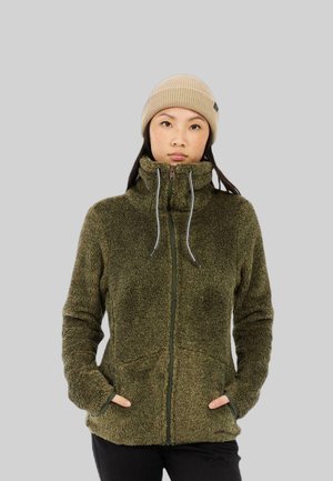 Mujer con un gorro beige y una chaqueta polar con cremallera y cuello alto de color verde oliva, con las manos en los bolsillos, de pie frente a un fondo gris.