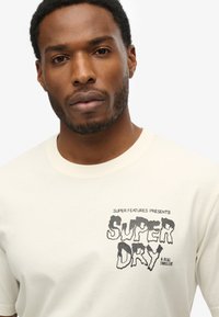 Crème-kleurig t-shirt van katoen met een zwarte grafische print met de tekst "SUPER DRY" en "SUPER FEATURES PRESENTS" op de voorkant.