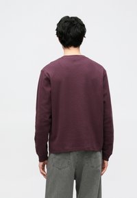 Bordowy długi sweter z długim rękawem, o prążkowanej fakturze, okrągły dekolt, luźny fason. Noszony z wyblakłymi szarymi spodniami o szerokich nogawkach, ręka oparta na boku.