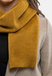 Scarf a maglia giallo senape con estremità a costine, che pende rilassatamente intorno al collo, abbinato a un maglione beige chiaro e un top nero.
