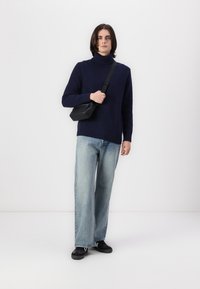 Navy gerippter Rollkragenpullover, kombiniert mit hellblauen, gerade geschnittenen Jeans, schwarzen Schuhen und einer kleinen schwarzen Umhängetasche. Einfarbiger Hintergrund.
