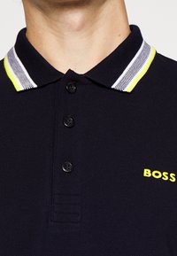 Polo shirt bleu marine avec un col classique, présentant des bordures rayées contrastantes en jaune et blanc, trois boutons noirs, et un logo jaune.