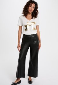 Witte V-hals t-shirt met gouden grafisch ontwerp, gecombineerd met zwarte leren broek met hoge taille en wijde pijpen. Zwarte platte schoenen maken de outfit compleet.